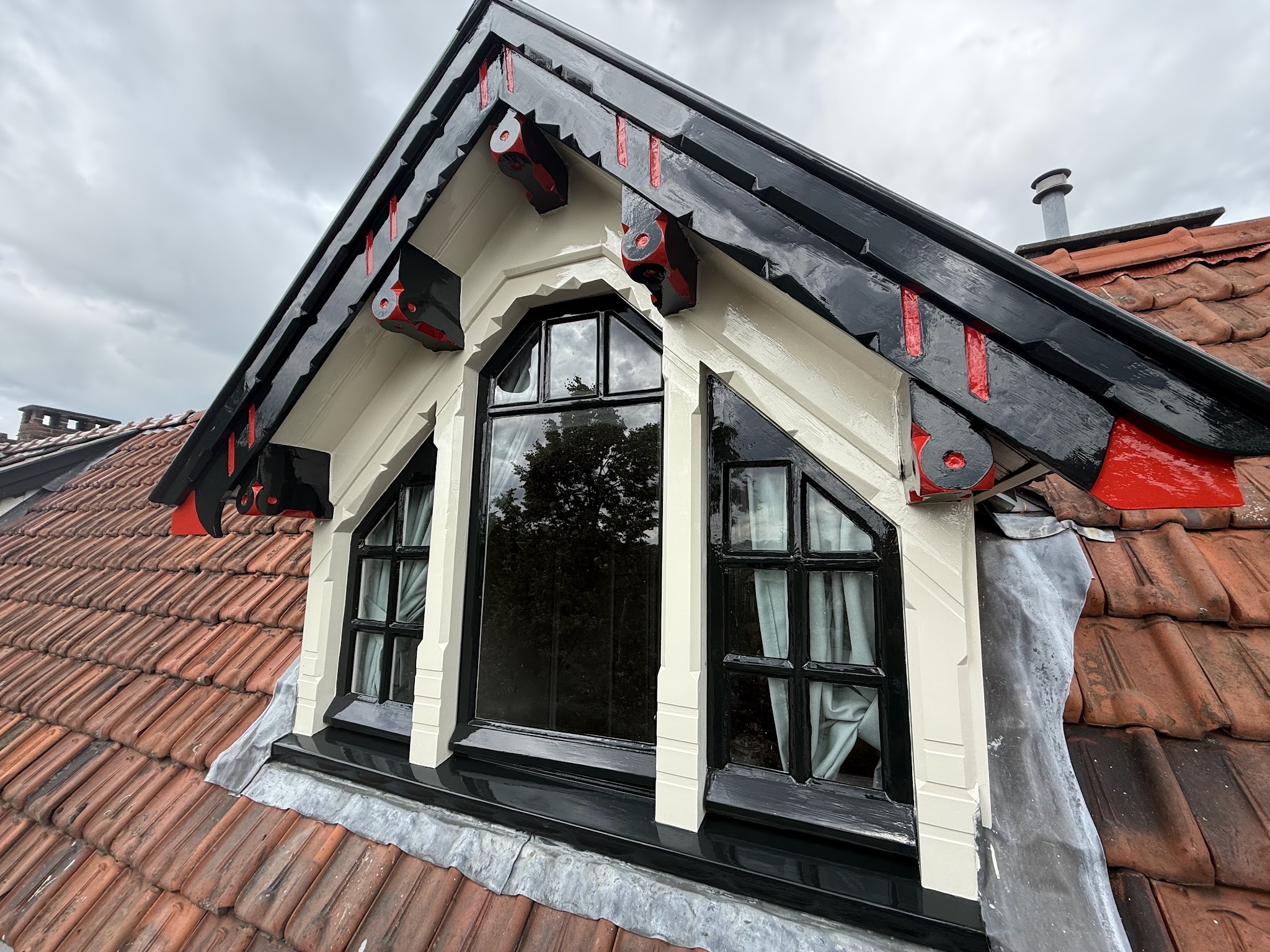 Dakkapel met crème kozijnen en zwart en rood geschilderd sierwerk op een pannendak